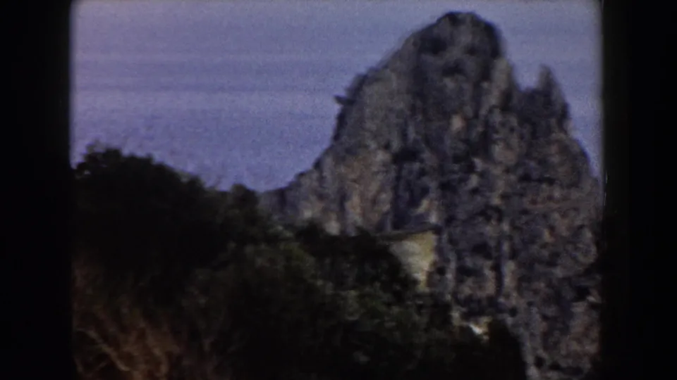 1960 Italy 8mm frame showing a tall weathered rock mass beside the sea, with dark vegetation at the base and soft blue-gray water behind it.