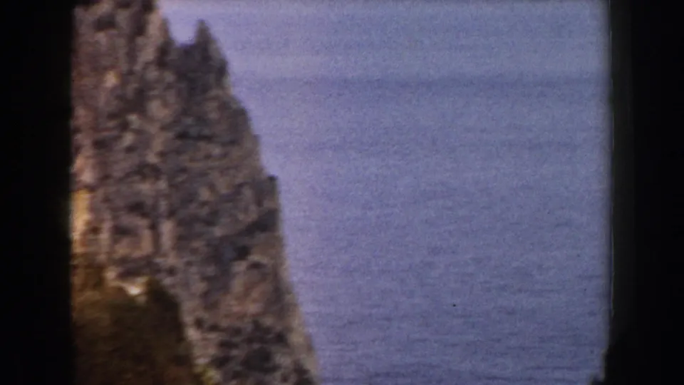 1960 Italy 8mm frame with a jagged coastal rock stack standing near calm blue-gray water, bordered by dark film edges.