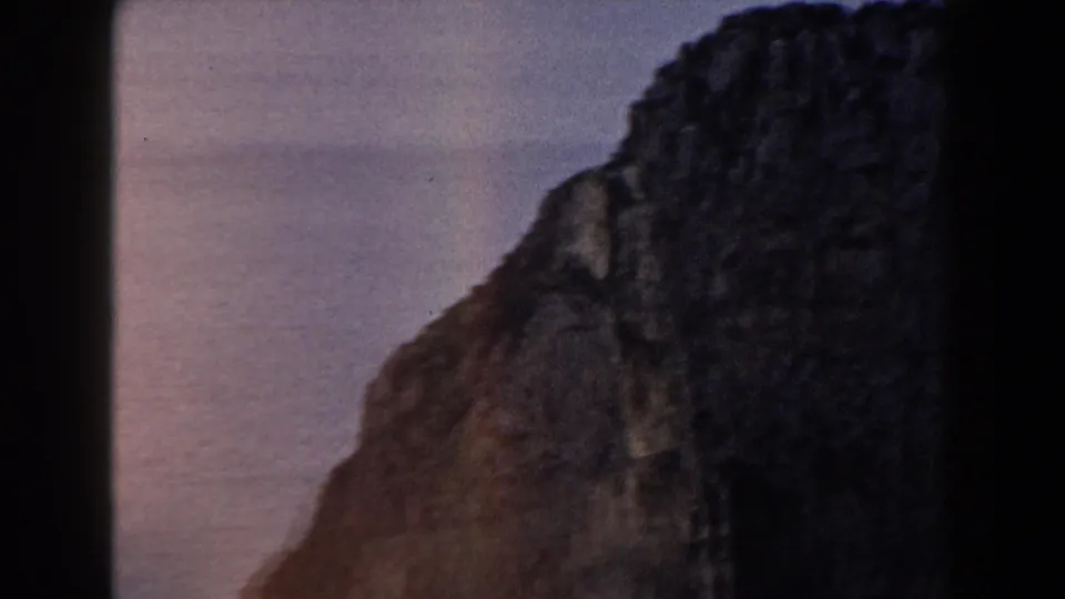 1960 Italy 8mm frame showing a broad, dark coastal rock face, with pale ocean and sky visible around its edges.