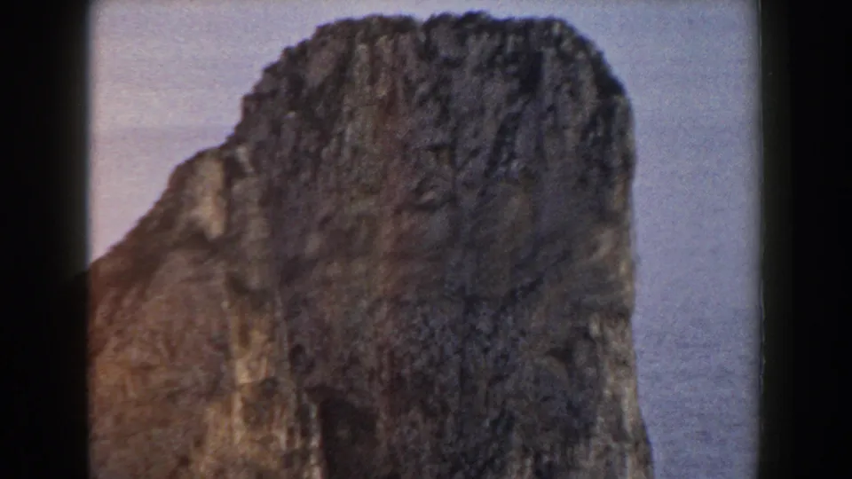 1960 Italy 8mm frame showing a broad weathered rock formation with textured tan-gray stone and calm sea behind it.