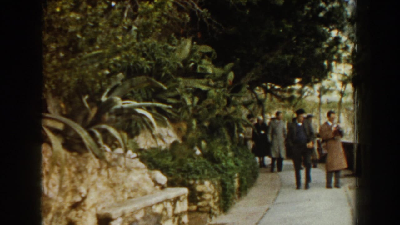 1960 Italy garden walkway with adults in period coats near stone borders, dense foliage, and agave-like plants.