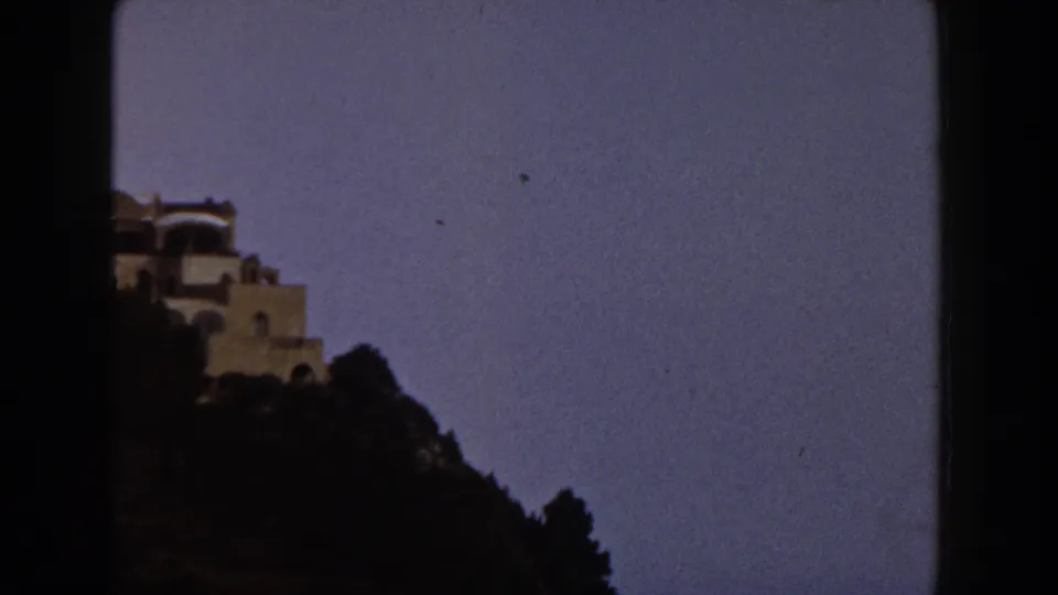 1960 Italy 8mm frame showing a pale Mediterranean-style villa on a hillside against a dusky blue sky.