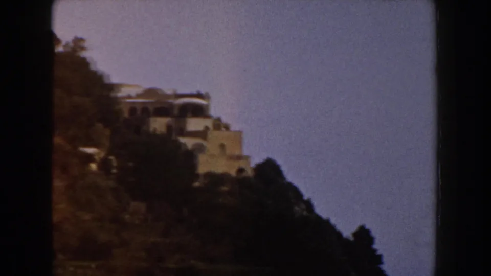 1960 Italy 8mm frame with a white hillside villa partly silhouetted above trees and rocky terrain.
