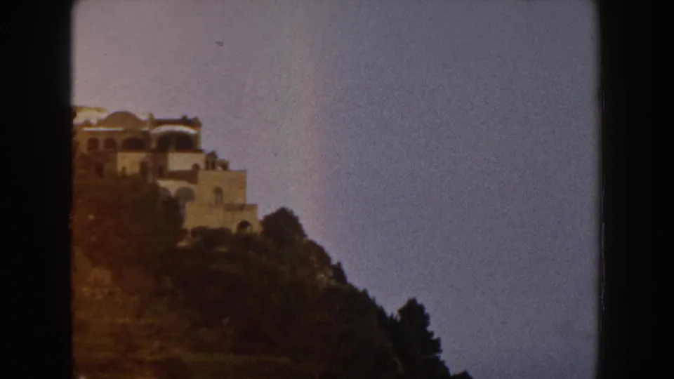 1960 Italy home-movie frame with pale hillside buildings partly cropped at left, trees below, and open blue sky.