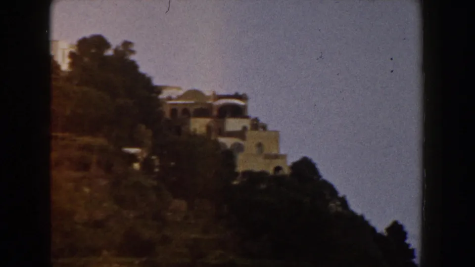 A 1960 Italy 8mm frame shows white hillside buildings, dark trees, brown slope, and blue sky with visible film borders.