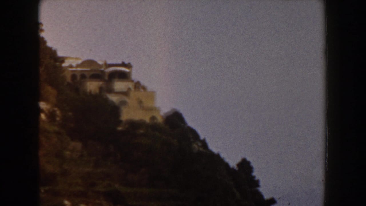 1960 Italy 8mm frame with a villa-like structure perched on a steep green slope, framed by sky and film gate edges.