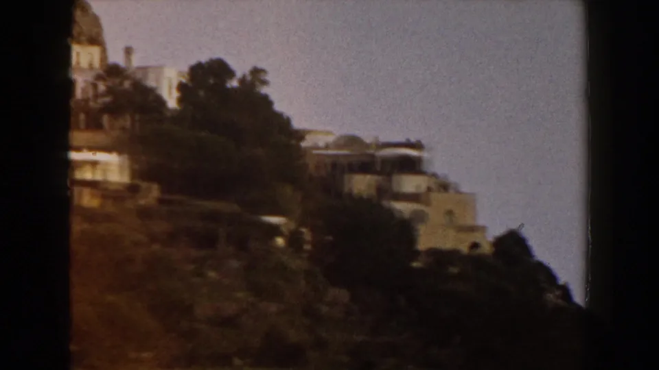 1960 Italy 8mm frame showing terraced hillside architecture partly obscured by trees, with the camera shifted lower on the slope.