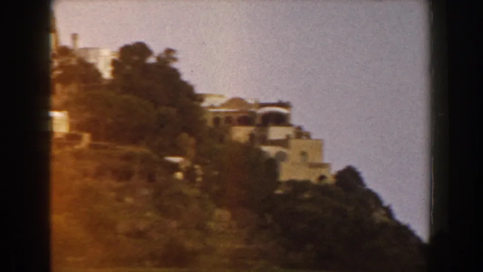 1960 Italy 8mm frame of a light hillside building among trees, with warm exposure, grain, and dark gate edges visible.