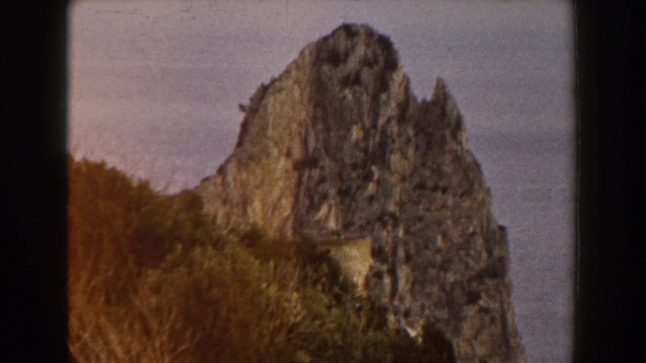 Jagged rocky cliff rising above brush with hazy open water or sky behind it.