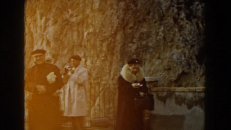 1960 Italy 8mm frame of adult tourists in coats and hats standing at a scenic overlook beneath a textured rock face.