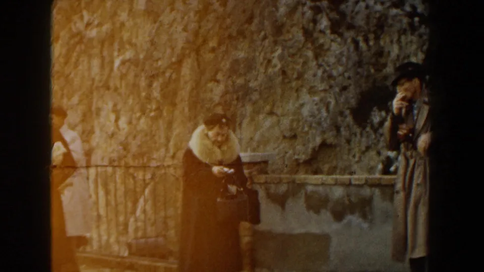 1960 Italy 8mm frame showing adults in mid-century coats at a rocky scenic overlook, with one person holding a camera near the wall.