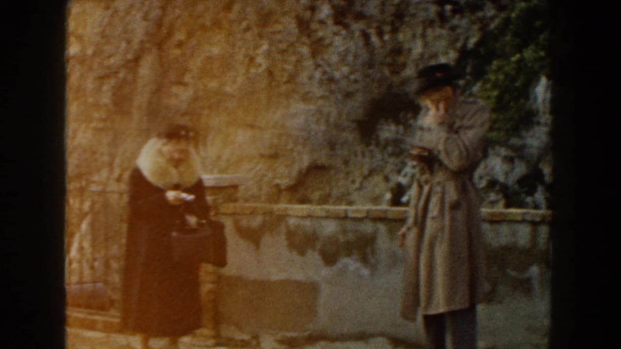 Two adults in coats and hats stand near a stone wall in bright outdoor light.