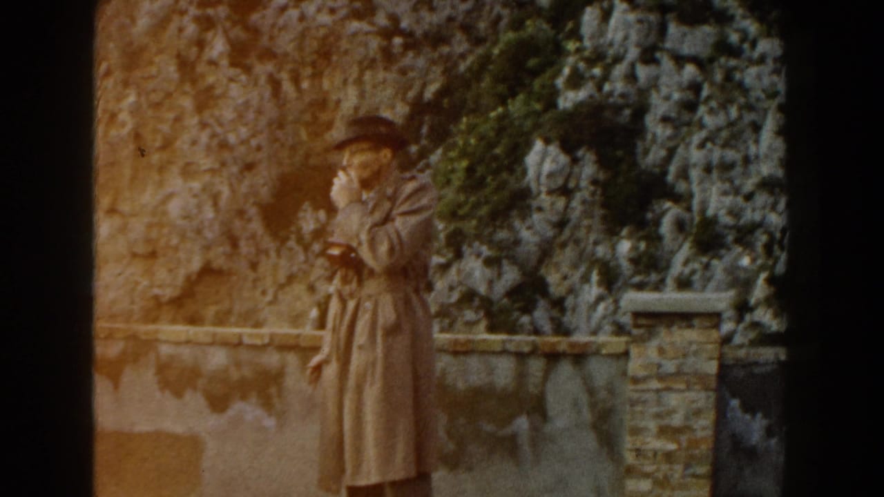 A person in a tan coat and hat stands alone by a stone wall and greenery.