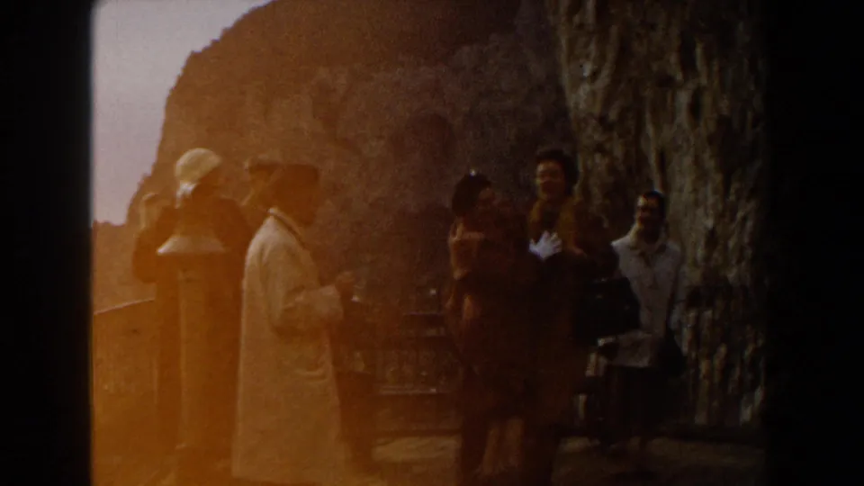 1960 Italy home-movie frame showing a small group of adults near a rocky wall, railing, and sun flare.