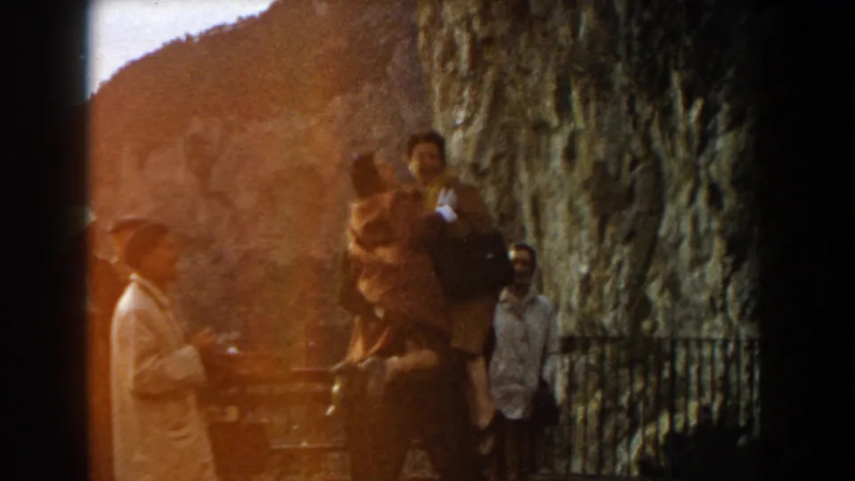 1960 Italy home-movie frame of adults beside a rugged rock face and railing, partly obscured by warm film flare.