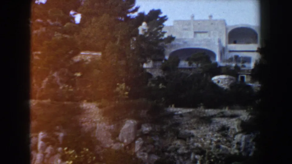 Wide 8mm view of a pale Italian hillside villa with arches, trees and rocky foreground.