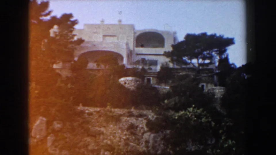 1960 Italy travel footage frames a white villa with arched openings above trees and rocks.