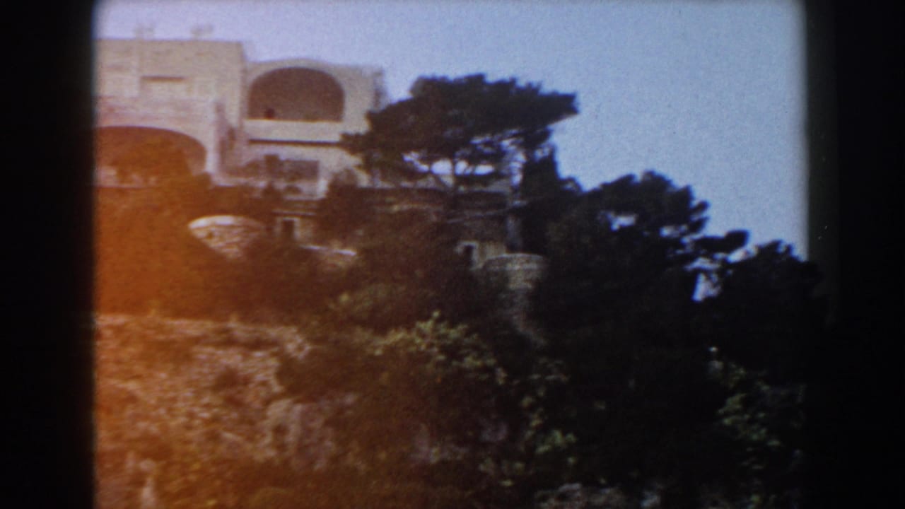 Pale hillside building above rocky terrain and dark trees.