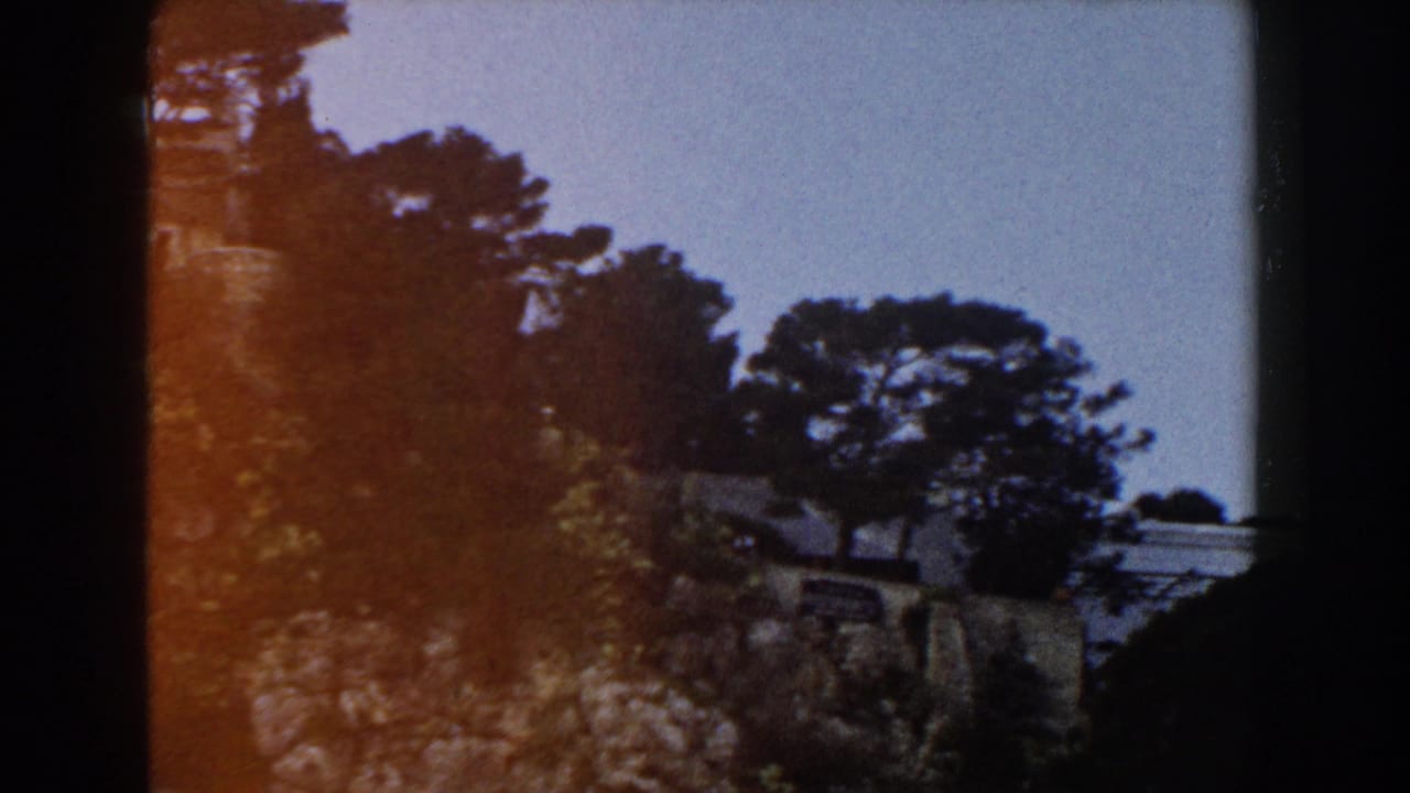 1960 Italy 8mm frame with trees silhouetted against sky, a rocky slope, and a pale hillside structure partly obscured.