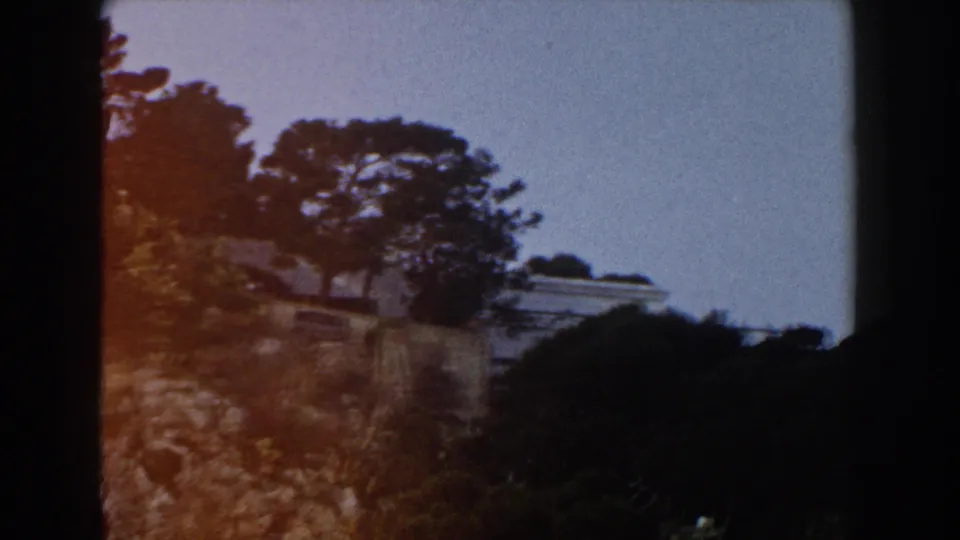 1960 Italy 8mm frame showing dark trees, rocky hillside plants, and a pale roofline or terrace beyond the slope.