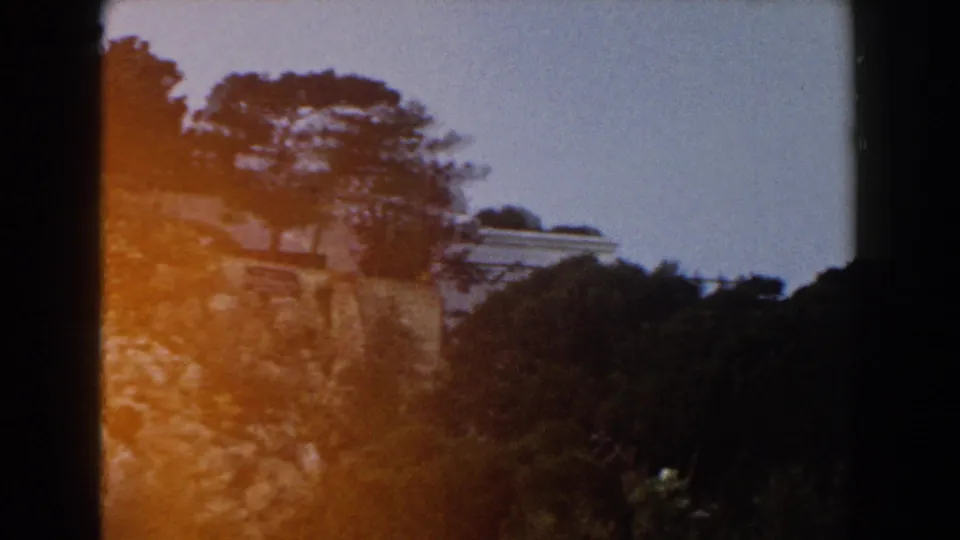 1960 Italy 8mm frame showing a rocky, tree-covered hillside with pale building edges visible beyond the foliage.