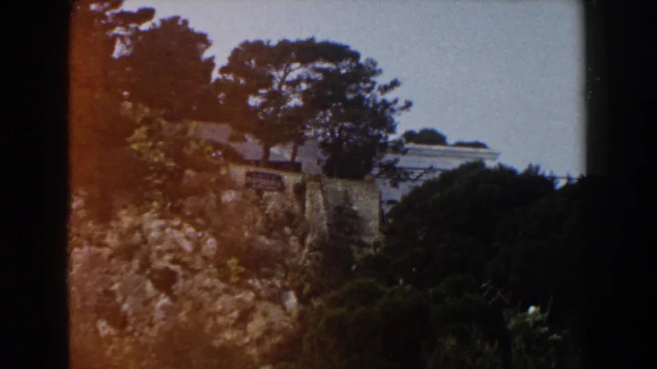 1960 Italy 8mm frame showing a rocky hillside, stone wall, tall trees, shrubs, and a pale building beyond the slope.