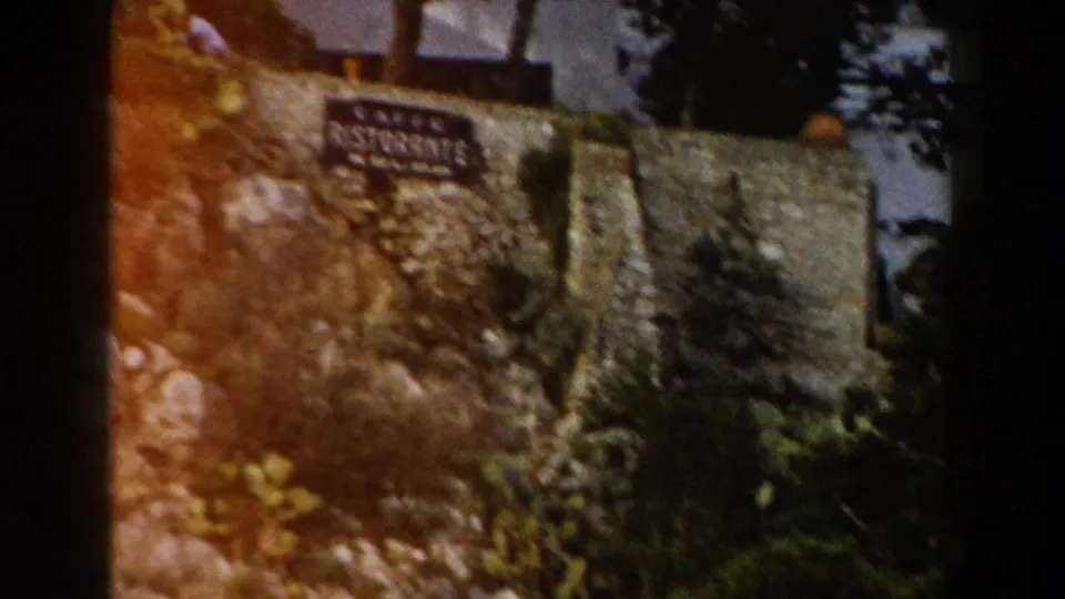 1960 Italy 8mm frame showing a Caffe Ristorante Krupp sign mounted on a rocky stone wall amid shrubs and trees.