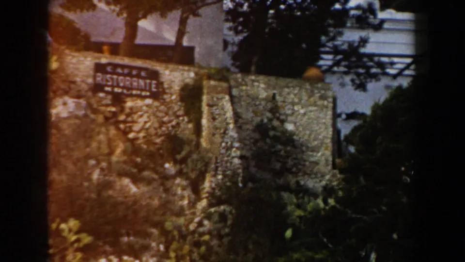 1960 Italy frame with the Caffe Ristorante Krupp sign, stone retaining wall, trees, shrubs, and white building details in view.