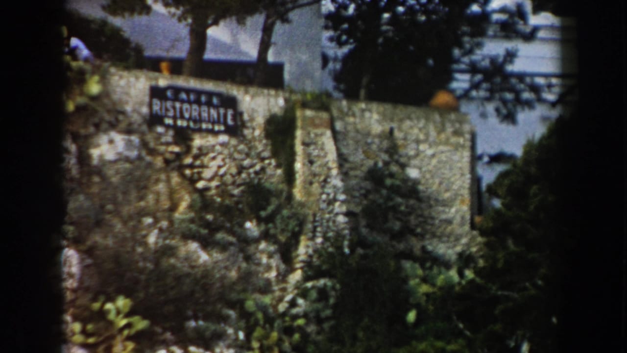 1960 Italy home-movie frame showing a roadside stone wall with a Caffe Ristorante sign, trees, and dense greenery.