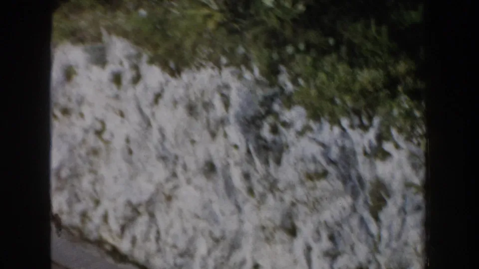 1960 Italy frame of a pale grey hillside rock face with patches of vegetation near the upper edge and visible 8mm border shadows.