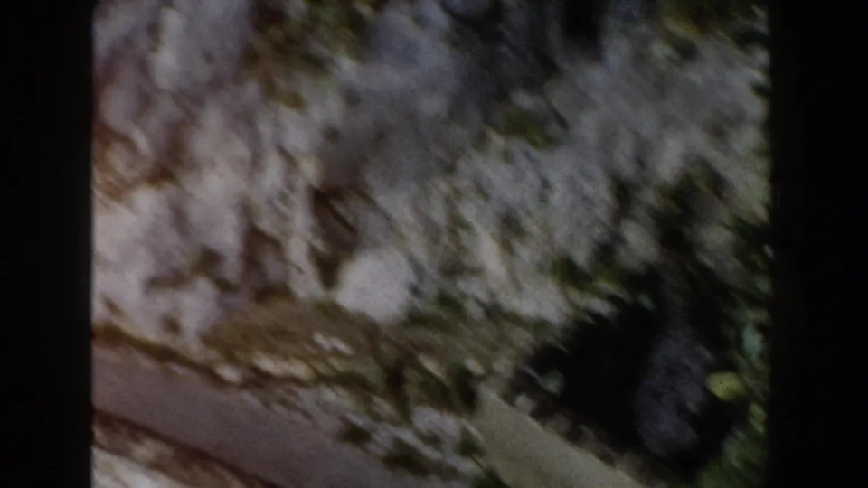 1960 Italy home-movie frame showing a rocky ledge with vegetation and a narrow road cutting along the lower edge.