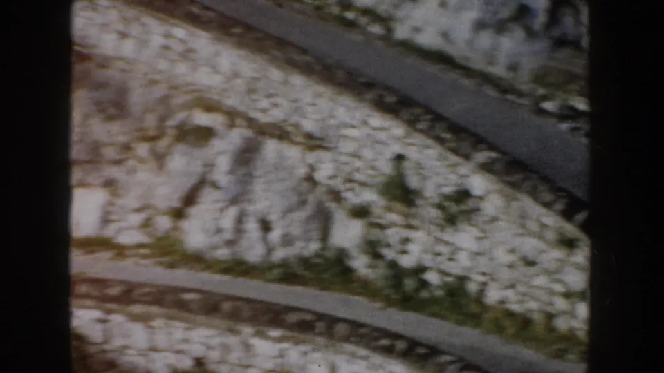 1960 Italy frame showing a winding hillside road bordered by pale stone and scattered vegetation, captured in soft 8mm color.