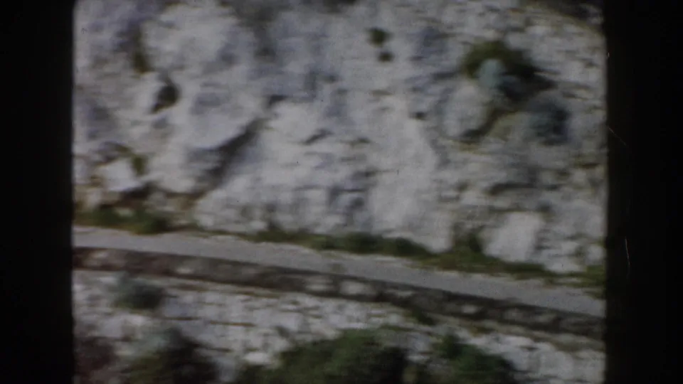 1960 Italy home-movie frame looking down on a narrow mountain road beside pale rock and sparse greenery.