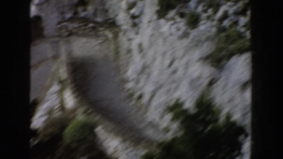 1960 Italy home-movie frame showing a blurred roadside edge, rocky slope, and green vegetation from above.