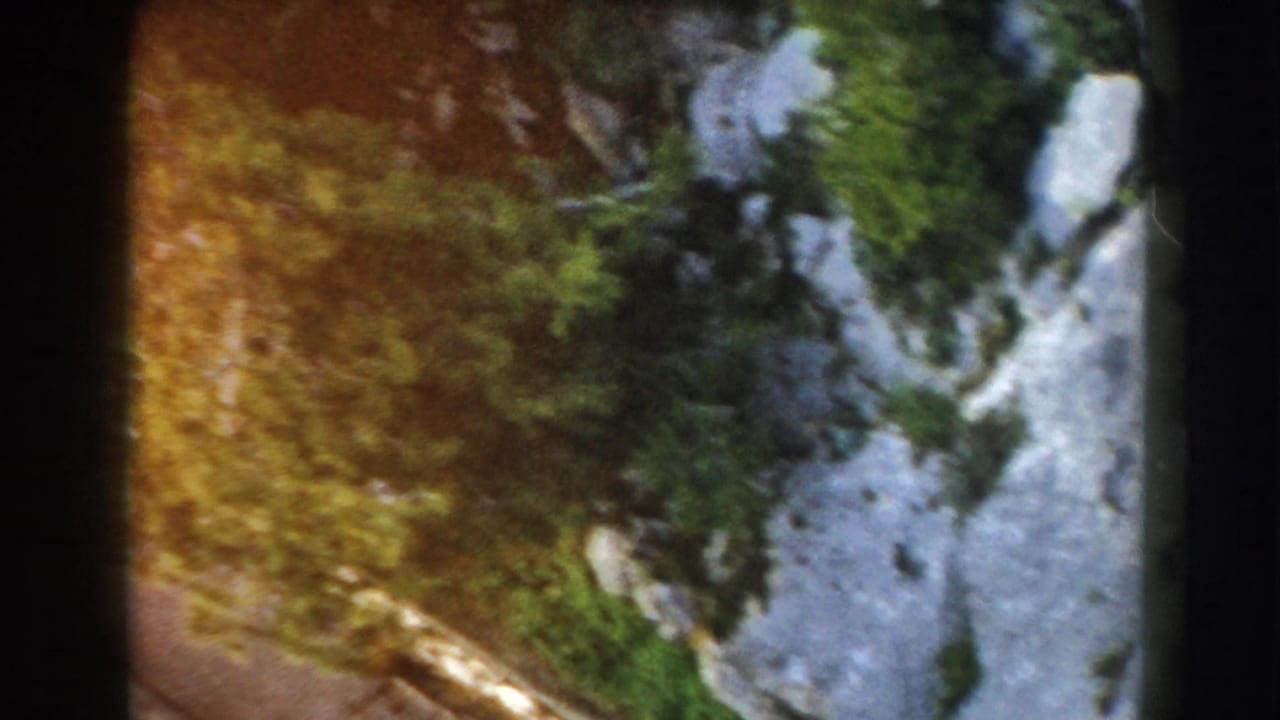 Steep rocky mountain terrain with trees and pale snow seen past dark film-frame edges.