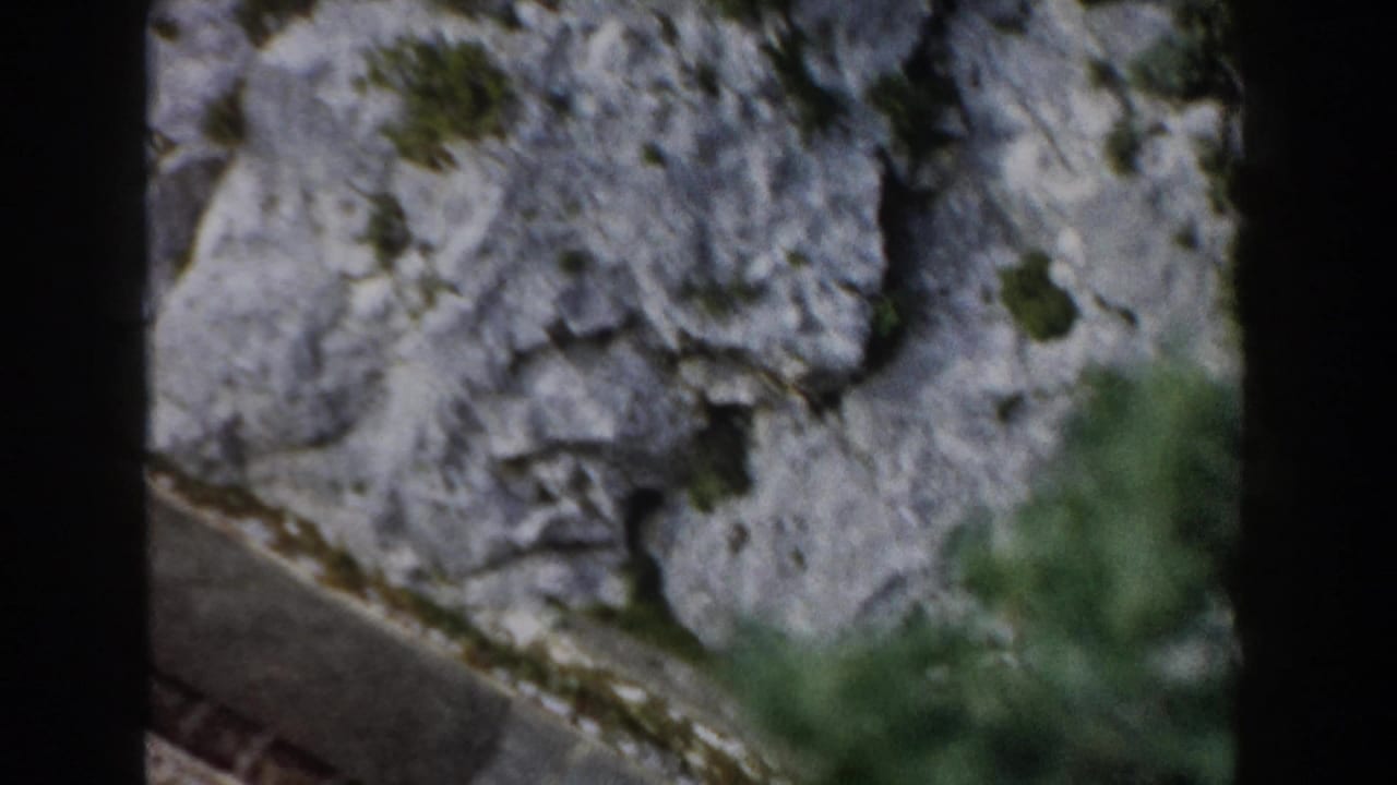 1960 Italy mountain view with a pale cliff face, a narrow road or track, and dark vertical framing from the moving camera position.