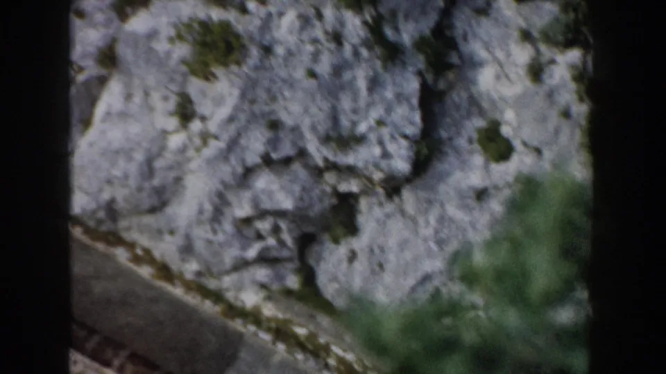 1960 Italy mountain view with a pale cliff face, a narrow road or track, and dark vertical framing from the moving camera position.