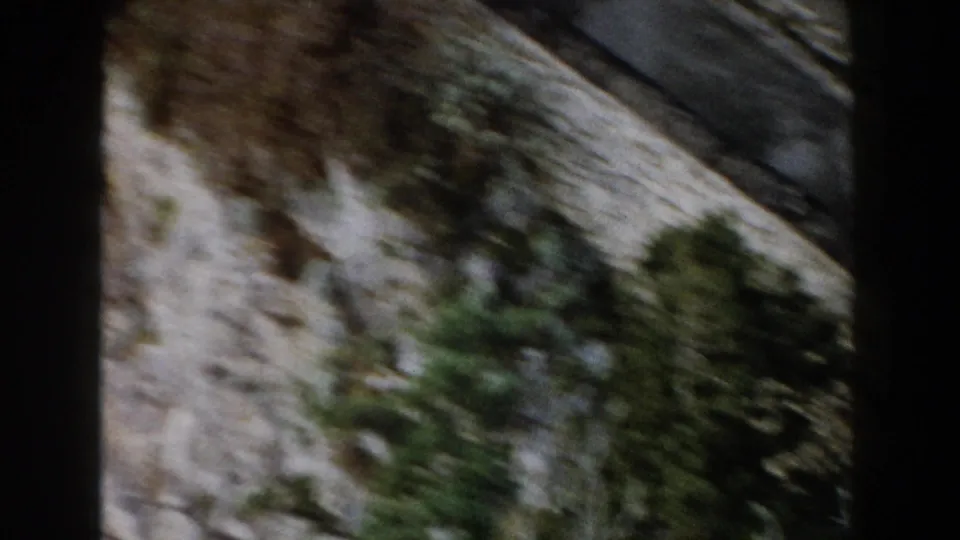 1960 Italy mountain-travel frame showing a diagonal road or track beside rocky slopes and scattered green trees.
