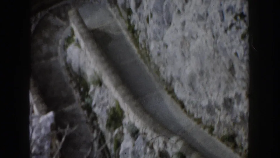 A high-angle 8mm view shows a winding paved edge running along a steep Italian rock face with small plants in crevices.