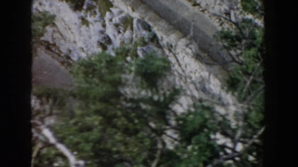 The frame shows a rocky Italian mountainside with a narrow paved line, shrubs, and dark tree branches crossing the lower view.