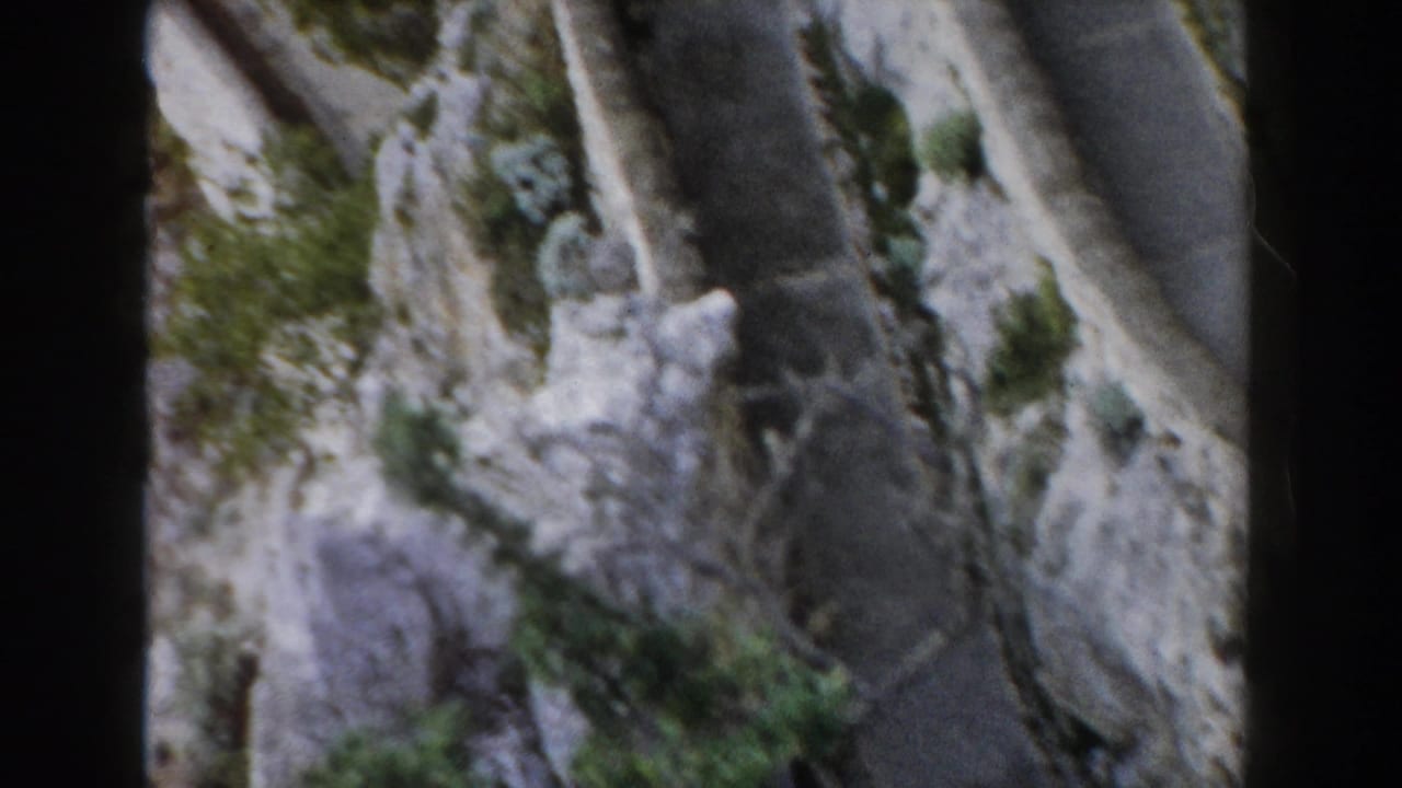 Curving mountain road beside pale rock and shrubs, seen from a moving vehicle.