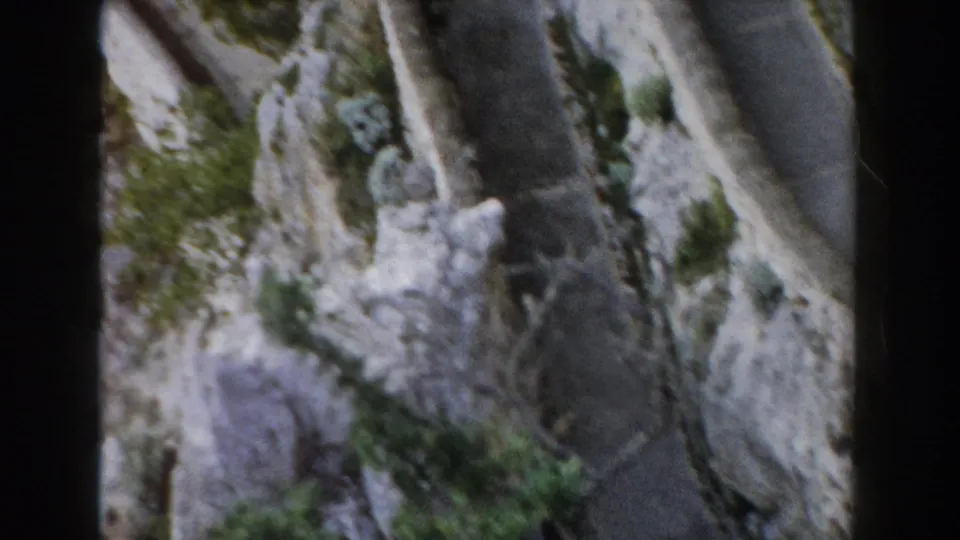 1960 Italian 8mm view of a narrow mountain road curving along rocky canyon terrain with sparse green vegetation.
