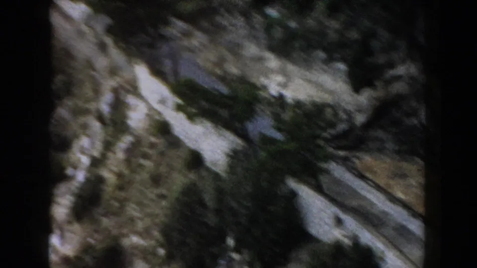 1960 home-movie frame from Italy showing a winding road through rugged rock and scrub vegetation from a vehicle perspective.