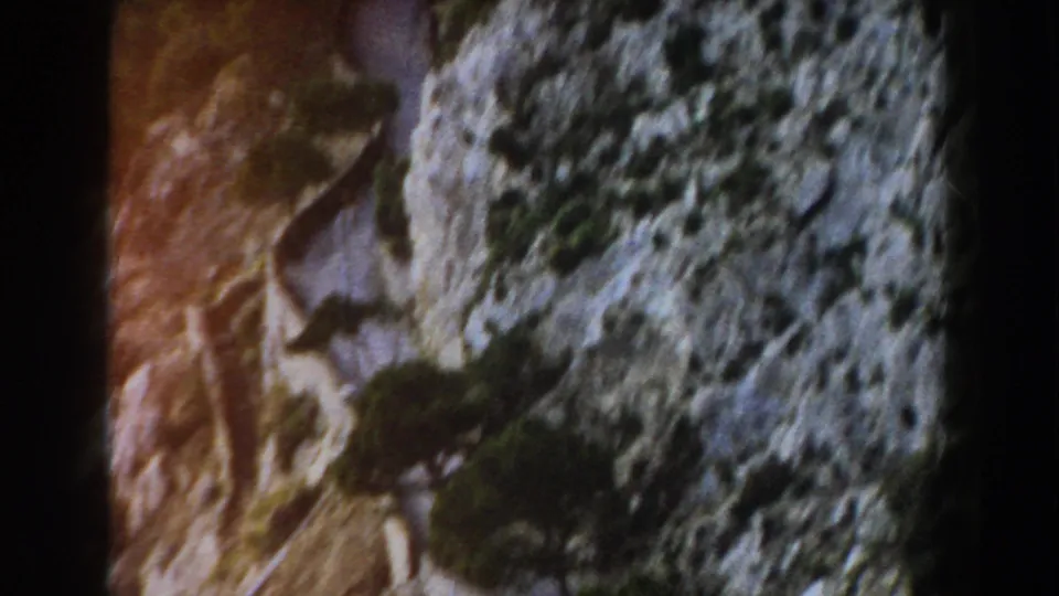 Italian 8mm travel footage frame showing a cliffside road bordered by pale stone, rough canyon texture, and sparse greenery.