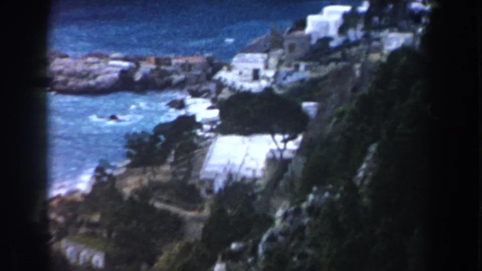 1960 Italy 8mm frame looking down toward blue sea, white coastal buildings, rocks, and a winding path.
