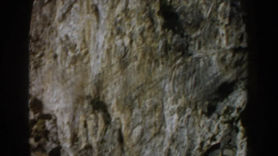 1960 Italy 8mm frame showing a mottled grey and green rock surface with small shadowed holes and uneven daylight exposure.