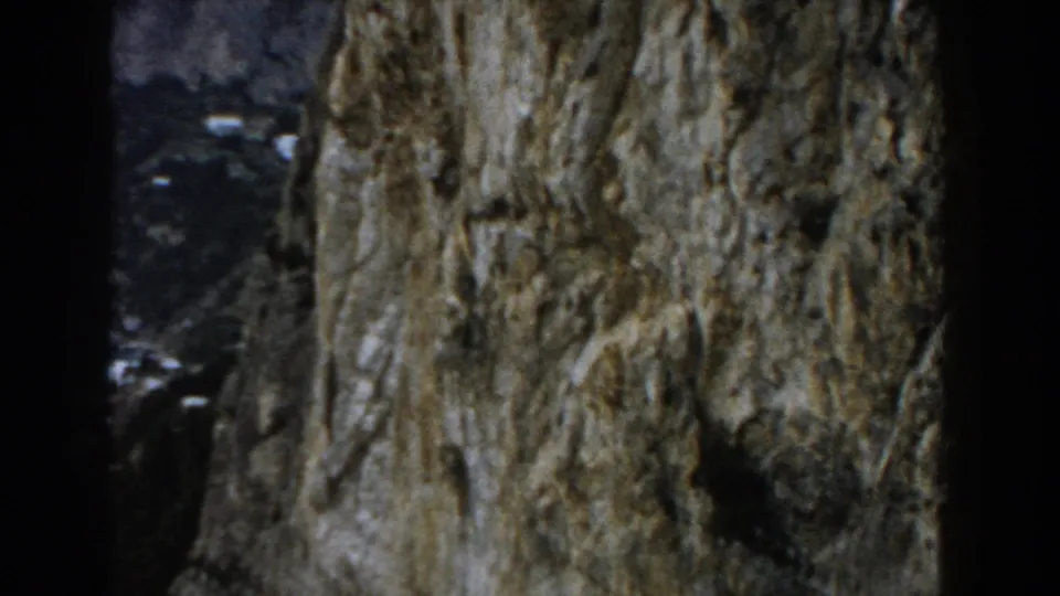 1960 Italy 8mm frame showing a rough cliff face close to camera with distant coastal hillside visible through dark edges.