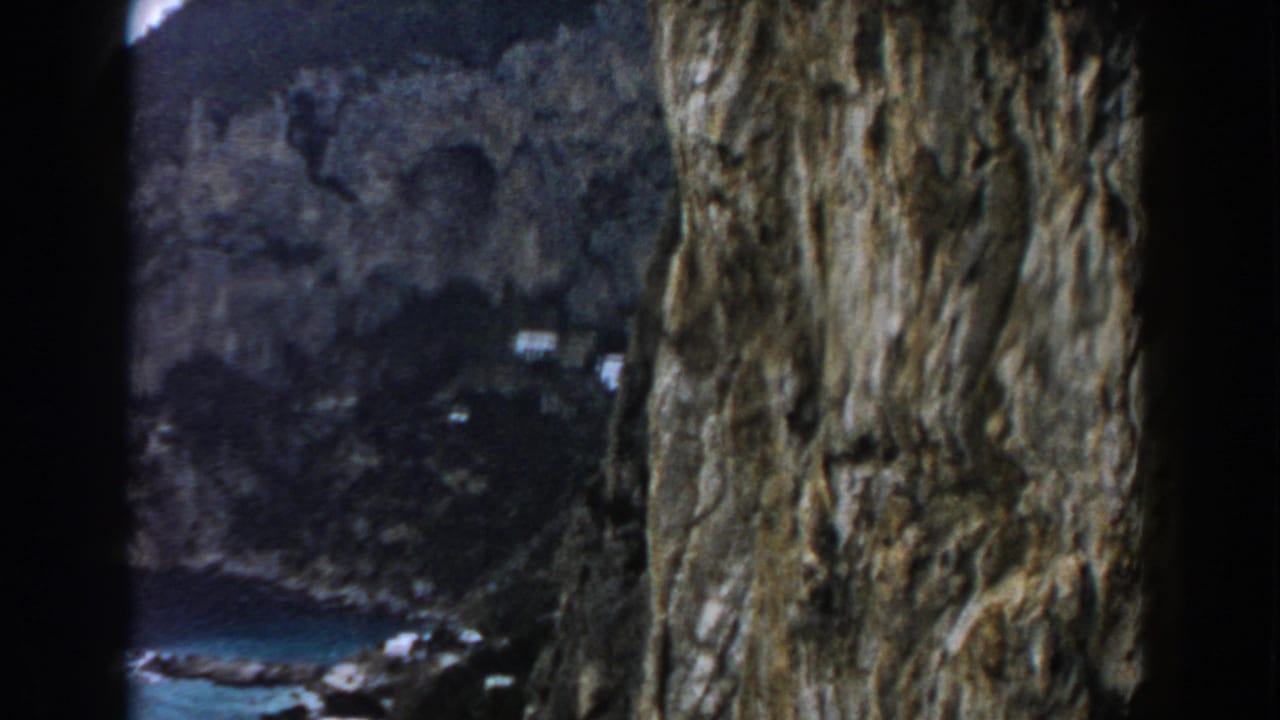 Cliff face beside a distant blue cove and shoreline buildings.