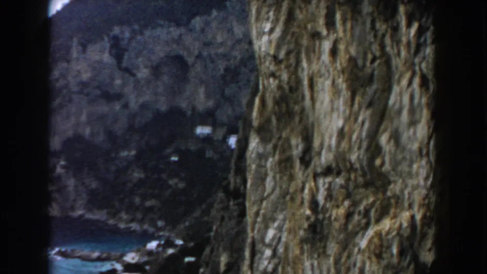 1960 Italy 8mm frame with a steep rock wall in the foreground and a blue sea cove with small distant buildings below.