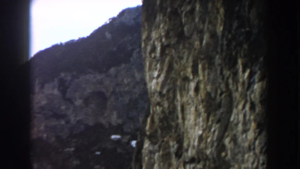 1960 Italy 8mm frame showing the cliff surface dominating the view, with a coastal hillside and small structures in the distance.
